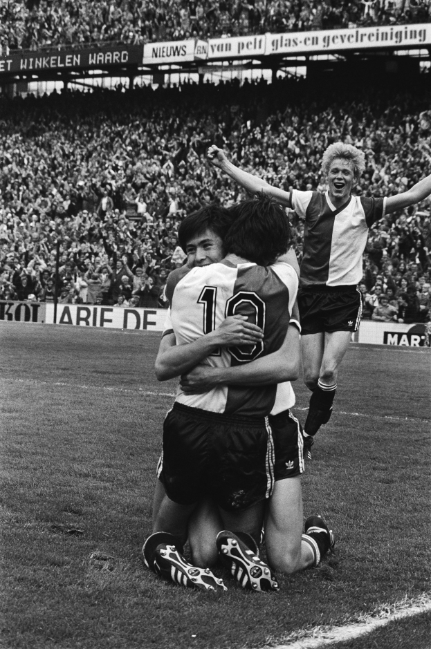 Drei Füßballspieler umarmen sich auf dem Boden nach einem Tor, mit jubelnder Menge und schwenkenden Fahnen in einem schwarz-weiß Stadion Setting.