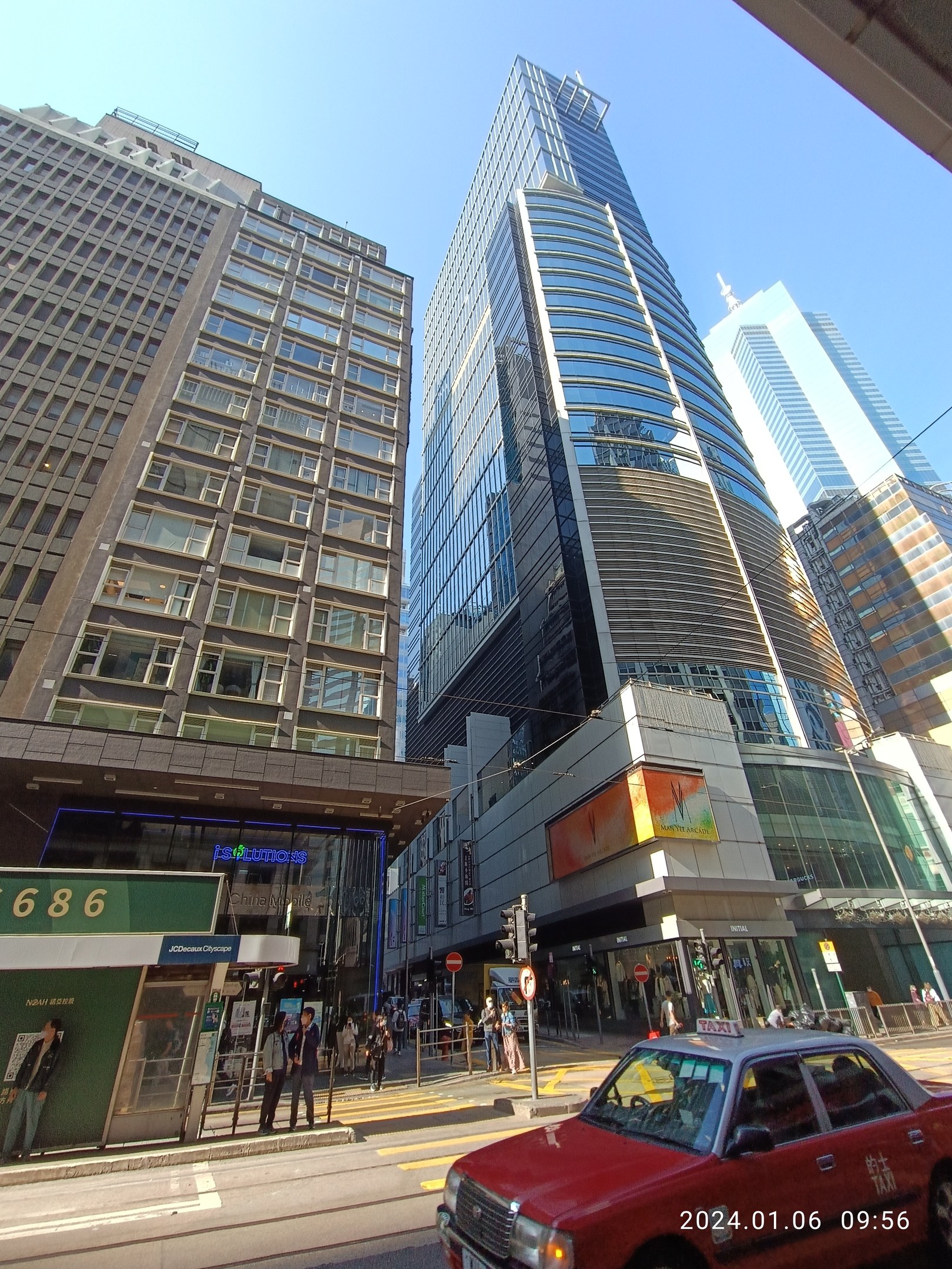 Roter Wagen auf einer von Hochhäusern gesäumten Stadtstraße, Fußgänger auf dem Bürgersteig, Verkehrsampeln, Schilder und Namenskarten, mit Himmel im Hintergrund.
