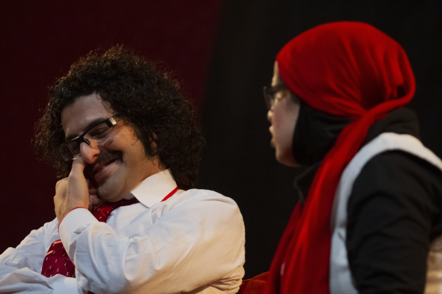 Ein Mann in weißem Hemd und roter Krawatte sitzt neben einer Frau mit Brille auf einer dunklen Bühne.