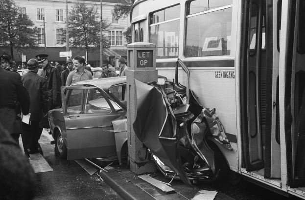 Schwarz-weißes Bild eines Busses, der auf der Seite der Straße in ein Auto gekracht ist, mit Menschen, die um die Szene stehen und Bäumen und Gebäuden im Hintergrund.