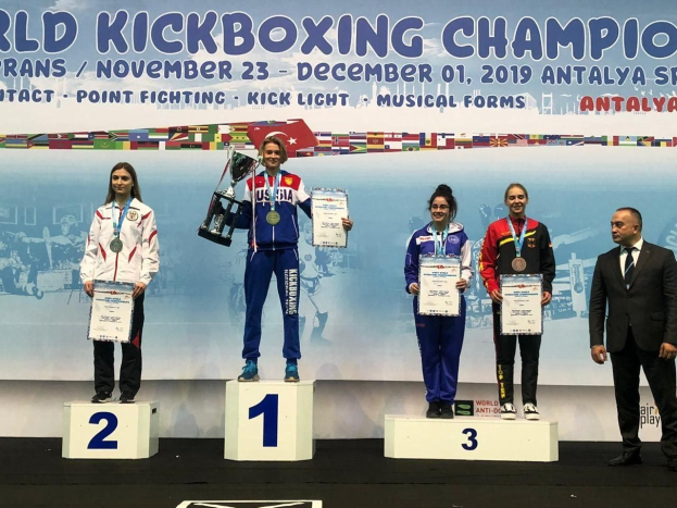 Fünf Personen auf einem Podium mit Trophäen und Zertifikaten, im Hintergrund ein Banner 'Welt-Kickbox-Meisterschaft Antalya 2019'