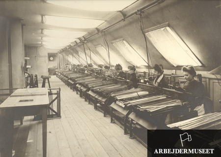 Schwarz-weiß-Foto von Frauen, die an Textilmaschinen in einer Fabrik arbeiten, die auf Stühlen sitzen und links von ihnen Tische stehen, eine Uhr an der Wand im Hintergrund und Text in der rechten unteren Ecke.