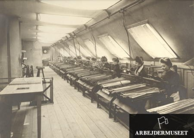 Schwarz-weiß-Foto von Frauen, die an Textilmaschinen in einer Fabrik arbeiten, die auf Stühlen sitzen und links von ihnen Tische stehen, eine Uhr an der Wand im Hintergrund und Text in der rechten unteren Ecke.