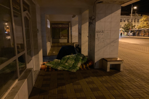 Eine Person schläft auf einer Decke und einer Bank auf einem Gehweg bei Nacht, mit einer Gruppe von Menschen in der Nähe, Fahrzeugen auf der Straße, einem Straßenpfahl, Bäumen, einem Schild, Gebäuden mit Fenstern und einem bewölkten Himmel.