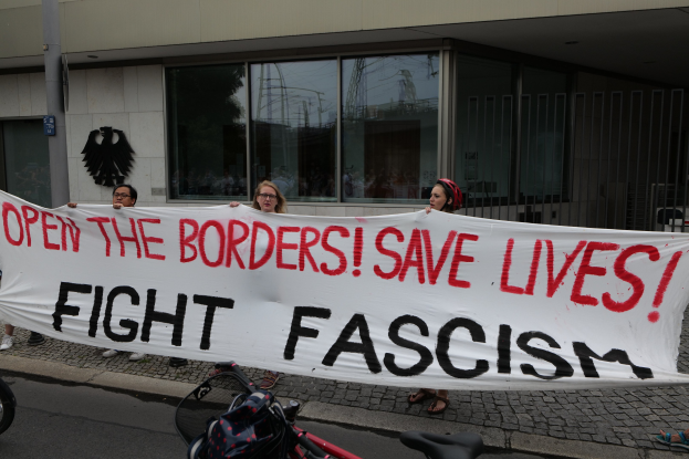 Eine Gruppe von Menschen hält eine Fahne mit der Aufschrift "Öffnet die Grenzen, rettet Leben, kämpft gegen Faschismus" vor einem Gebäude mit Glasfenstern und einem Mast, mit einem Fahrrad und einer Tasche auf der Straße.