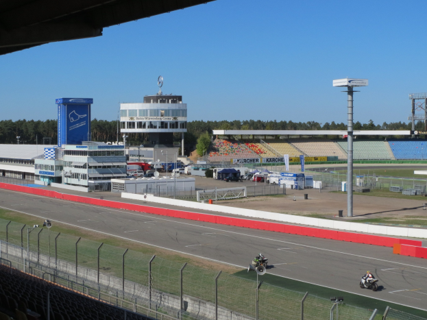 Gruppe von Motorradfahrern auf einer Rennstrecke, umgeben von einem Metallzaun, Gras, Fahrzeugen, einem Gebäude, einem Schild, Pfählen, Stadion, Deckenleuchten, Bäumen und einem bewölkten Himmel.