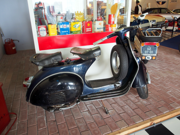 Ein schwarzer und weißer Roller steht vor einer Tankstelle, mit einem roten Objekt und einem Feuerlöscher links daneben und mehreren Fahrzeugen, einer Person und Brettern an der Wand im Hintergrund.