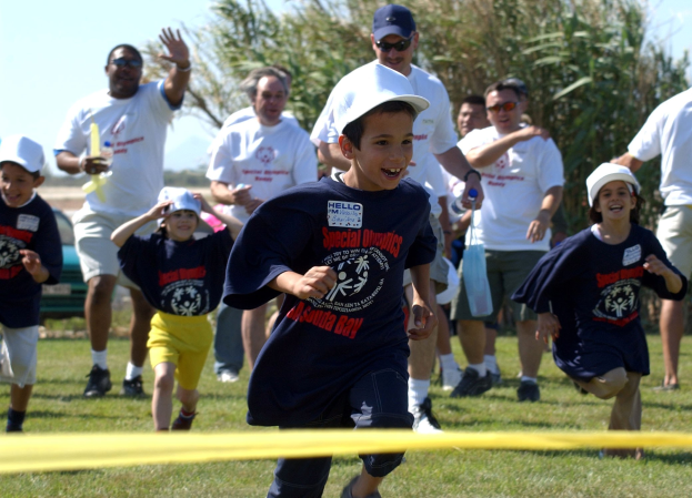 Gruppe von Kindern bei einem Lauf auf einer grünen Wiese mit einem gelben Band im Vordergrund, einige tragen Mützen und Schutzbrillen, andere halten Gegenstände, mit Bäumen und einem klaren blauen Himmel im Hintergrund.