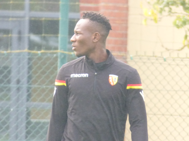 Ein Mann In Einer Schwarzen Jacke Steht Auf Einem Fussballfeld, Umgeben Von Einem Zaun Und Einem Baum, Scheint Konzentriert Während Einer Trainingseinheit.