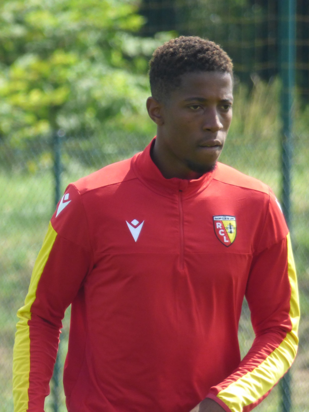 Junger Mann in rotem und gelbem Fußballtrikot auf einem Feld mit einem Zaun und Bäumen dahinter, der konzentriert wirkt.