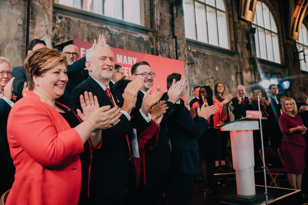 Eine Gruppe von Menschen, die vor einem Publikum stehen und jubeln, mit einem Podium, einem Mikrofon und einer Tafel mit Text auf der rechten Seite sowie Stühlen, einer Fahne, einer Wand, Fenstern und Lichtern im Hintergrund.