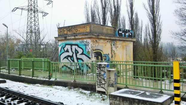 Ein Zuggleis mit Graffiti an der Seite, umgeben von Geländern, Pfählen, Lichtern und Bäumen, mit Türmen und Drähten im Hintergrund und Schnee am Boden.