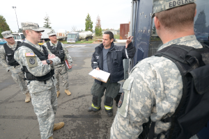 Eine Gruppe von Menschen in Uniformen, Mützen, Jacken und Schuhen steht auf einer Straße, wobei eine Person in einer schwarzen Jacke ein Blatt mit Papieren und einen Stift in der Nähe eines Fahrzeugs hält; im Hintergrund sind weitere Fahrzeuge, Bäume, Gras, ein Zaun, ein Lichtmast und ein klarer Himmel zu sehen.