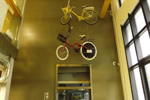 Zwei Fahrräder hängen von der Decke, ein Glasfenster auf der rechten Seite und ein Metallrohr auf der linken Seite in einem Gebäudeeingang.