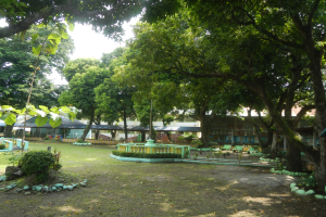 Ein Park mit saftigem Grün, Pflanzen und Bäumen, der eine zentrale Fontäne und Gebäude sowie einen klaren blauen Himmel im Hintergrund zeigt.