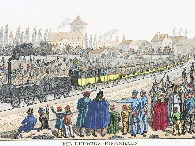 Eine Gruppe von Menschen steht in der Nähe eines Zuges auf Schienen, einige sitzen im Zug und andere auf dem Boden, vor einem Hintergrund aus Gebäuden und Bäumen, mit Text unten, der "Ludwigs-Eisenbahn, der erste Bahnhof in Deutschland" lautet.