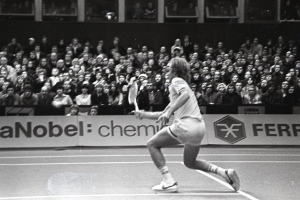 Schwarzes Bild einer Frau, die auf einem Tennisplatz spielt, mit Zuschauern auf den Rängen und einem Banner im Hintergrund.