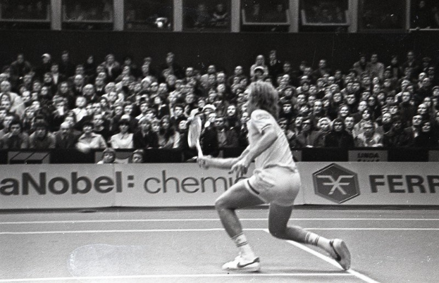 Schwarzes Bild einer Frau, die auf einem Tennisplatz spielt, mit Zuschauern auf den Rängen und einem Banner im Hintergrund.