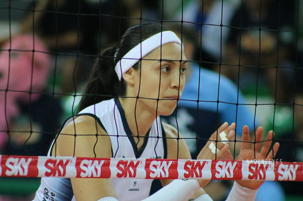 Eine Volleyballerin in Aktion auf dem Feld, mit Netz im Vordergrund und unscharfem Hintergrund, zeigt eine entschlossene Miene.
