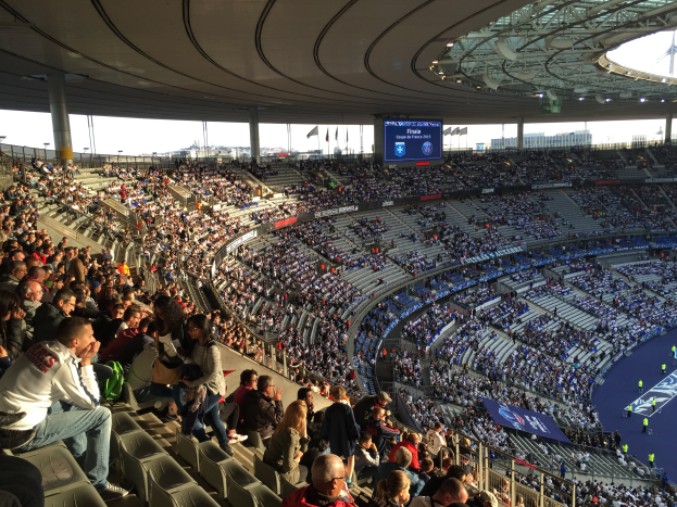 Große Menschenmenge in einem Stadion, das ein Förspiel schaut, mit einer Bühne auf der rechten Seite, Fahnen, Masten, einem Bildschirm und der Allianz Arena in München, Deutschland im Hintergrund.