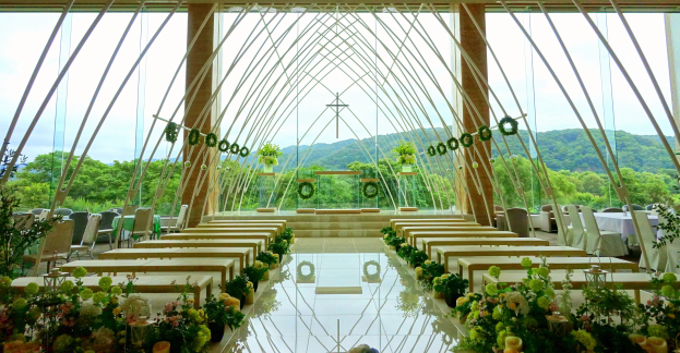 Eine Hochzeitsfeier in einem großen Saal mit Tischen in Halbkreisform, dekoriert mit Blumen, vor einer Kulisse aus Bäumen, Hügeln und einem klaren Himmel.