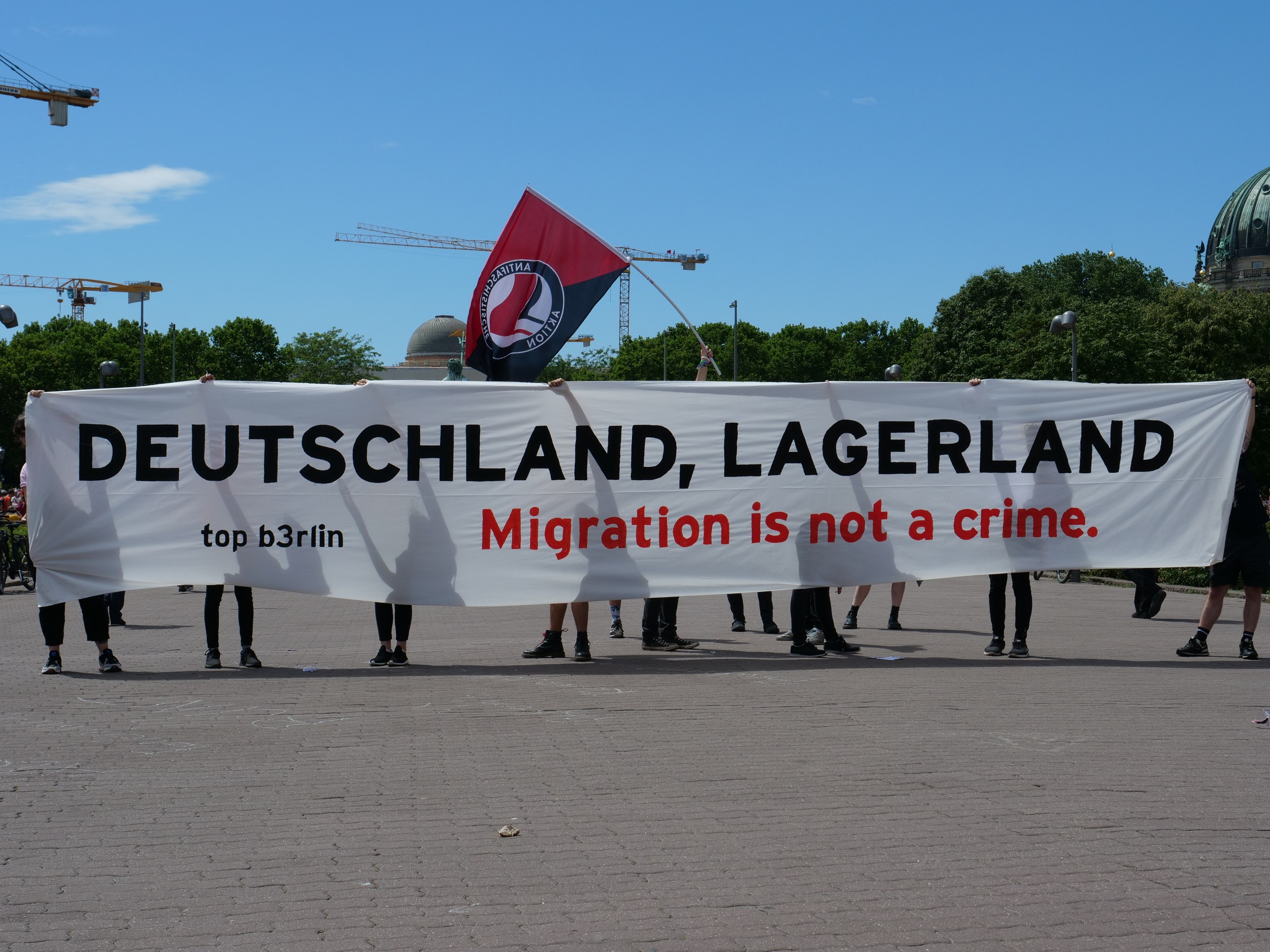 Gruppe von Menschen mit einem Banner 'Deutschland, Lagerland Migration ist kein Verbrechen' und einer Flagge, mit Straßeninfrastruktur, Bäumen, einem Gebäude und einem bewölkten Himmel im Hintergrund.