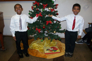 Ein geschmückter Weihnachtsbaum steht in einem Raum mit einer Kiste und anderen Gegenständen auf dem Boden, einem hölzernen Tisch links, einem Stuhl rechts und einer weißen Wand im Hintergrund mit einigen Gegenständen daran befestigt. Zwei Jungen in weißen Hemden lächeln und halten den Weihnachtsbaum, wobei die Beine einer Person auf der rechten Seite des Bildes sichtbar sind.