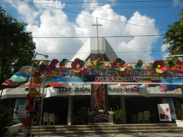 Ein Gebäude mit einer Tafel, auf der "Frohes Fest St. Josef der Arbeiter" steht, mit einer Treppe, einem gewölbten Eingang mit Verzierungen, Außenbestuhlung, Topfpflanzen, Bäumen und einem bewölkten Himmel.