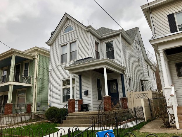 Reihe von Häusern mit Fenstern, Türen, Säulen, Geländern, Treppen und Dächern, umgeben von einem Metallzaun, Gras, Pflanzen, Bäumen und einem bewölkten Himmel, mit einem Mietschild am Boden, das die Adresse und Listen Details anzeigt.