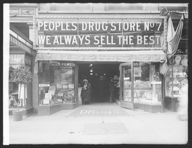 Ein Schwarz-Weiß-Foto eines Drugstores mit einem Schild, auf dem "People's Drug Store No. 7 - Wir verkaufen immer das Beste" steht, mit ein paar Menschen drinnen und einem Topf mit einer Pflanze links daneben.