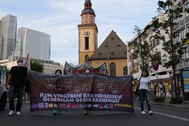 Eine Gruppe von Menschen mit Masken geht eine Straße entlang und hält ein Banner hoch, mit einem geparkten Auto auf der linken Seite, Gebäuden, Bäumen, Schildern, Pfosten und einem Uhrenturm im Hintergrund unter einem klaren blauen Himmel.