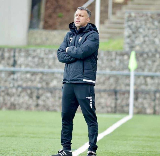 Ein Fußballtrainer steht auf einem Feld mit verschränkten Armen, trägt eine schwarze Jacke und Hose, vor einem Zaun, Treppe und Wand während eines Trainings.