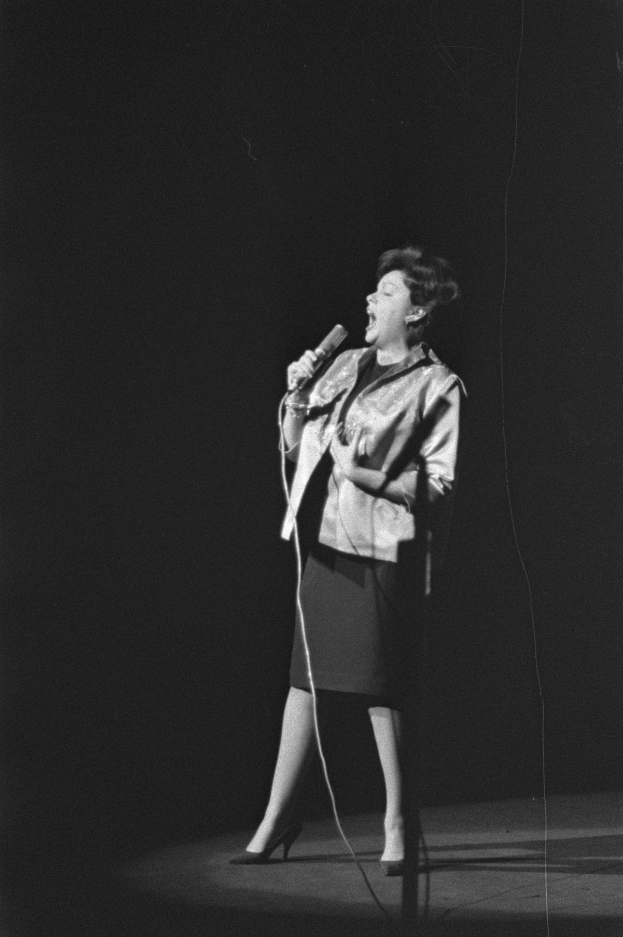 Eine Frau in einem schwarzen und weißen Outfit aus den 1950er-Jahren singt fröhlich auf einer von einem Scheinwerfer beleuchteten Bühne in ein Mikrofon, ihr Haar ist im klassischen 1950er-Jahre-Stil frisiert.