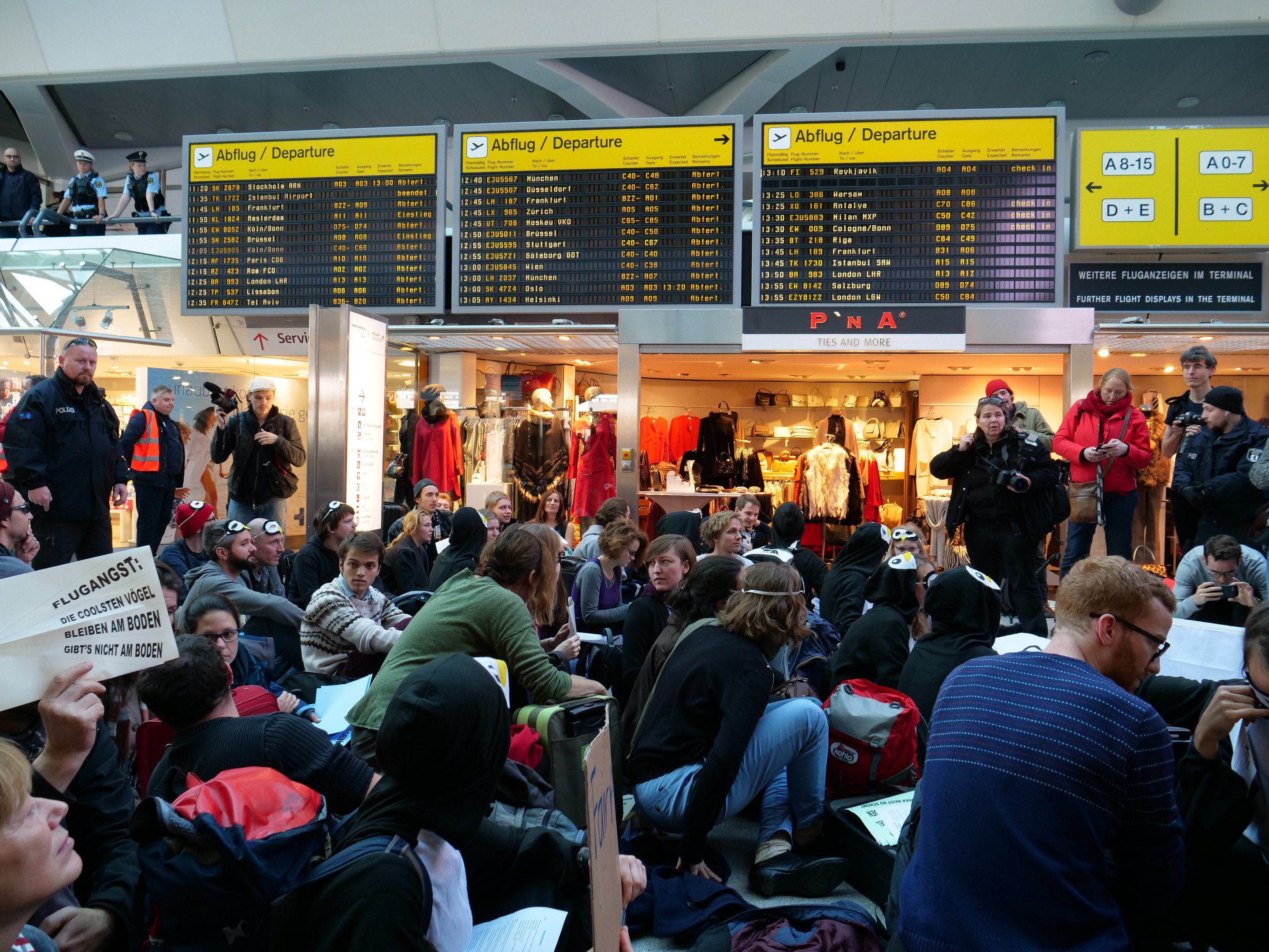 Große Gruppe von Menschen sitzt und steht in einem Flughafen-Terminal, einige halten Taschen und Papiere, mit Texttafeln und Schaufensterpuppen im Hintergrund, unter Deckenbeleuchtung, was auf eine Protestveranstaltung hindeutet.