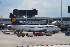 Ein Lufthansa Airbus A320-200 steht am Frankfurter Flughafen mit Fahrzeugen und Wagen auf der Straße davor, einem Gebäude, Laternenmasten und einem klaren blauen Himmel im Hintergrund.