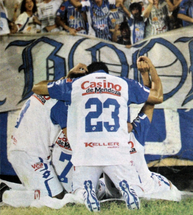 Gruppe von Fußballspielern, die auf dem Boden vor einer jubelnden Menge feiern, mit einem Banner im Hintergrund.