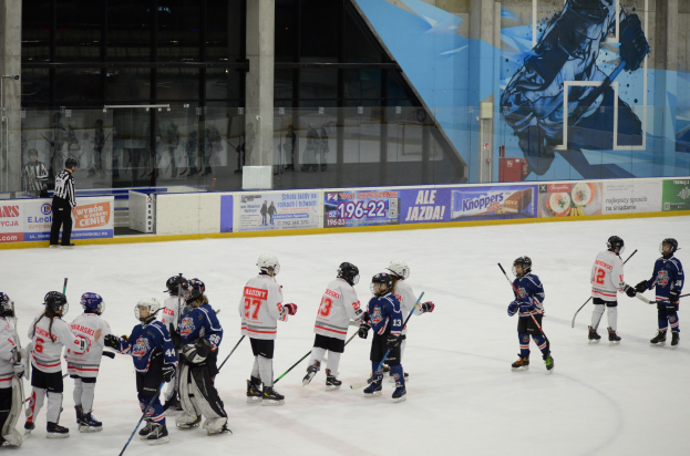 Gruppe junger Menschen beim Eisschlittschuhlaufen auf einer Halle, mit Helmen und Stöcken, mit einem Zuschauer links und Bannern, Säulen und Glaswänden im Hintergrund.
