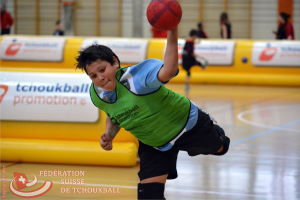 Ein Junge in einem weißen Hemd und dunklen Hosen wirft einen Ball mit seiner linken Hand, mit einem gelben Luftballon und mehreren Menschen im Hintergrund.