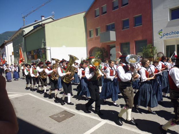 Eine Gruppe von Menschen in traditioneller bayrischer Tracht spielt Instrumente und trägt Fahnen, während sie eine Straße mit Gebäuden, Pflanzen und einem Schild entlanggehen, mit einem Hügel und einem blauen Himmel im Hintergrund.