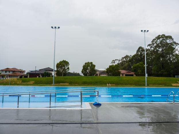 Große rechteckige Schwimmbad im Park, umgeben von Geländern, Pfosten und Lichtern, mit Bäumen, Häusern und einem klaren blauen Himmel im Hintergrund.