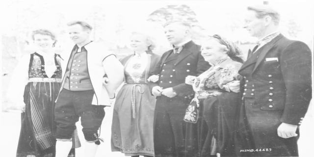 Eine Gruppe von Menschen in den 1930er Jahren, die in historischer Kleidung posieren, stehen zusammen im Freien mit Bäumen im Hintergrund und lächeln in die Kamera, mit dem Text "Bayerische Familie in den 1930er Jahren" unten auf dem Bild.