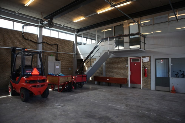 Ein großes Lagerinnere mit einem roten Gabelstapler, einem Wagen mit Gegenständen, einer Bank, einem Feuerlöscher, einer Treppe mit Geländern, Türen, Fenstern, Rohren und einem sichtbaren Rad.