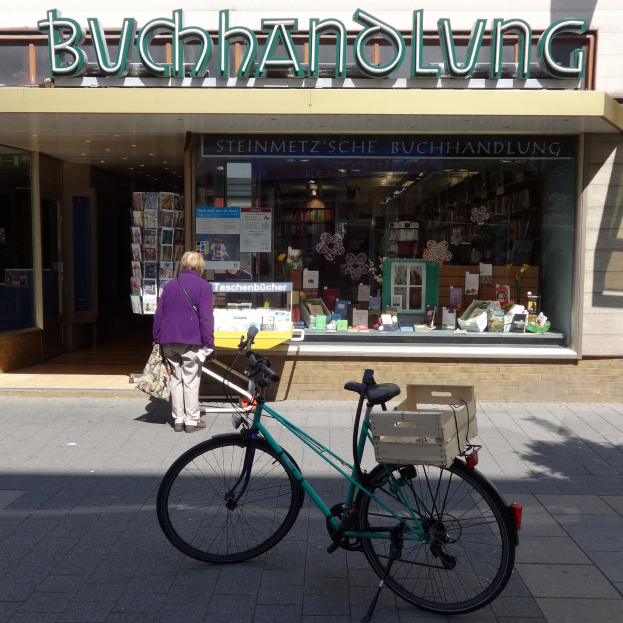 Eine Frau steht neben einem Fahrrad, das vor einem Geschäft geparkt ist, mit einem Fahrradrad im Vordergrund und Büchern, die durch das Schaufenster des Geschäfts sichtbar sind.