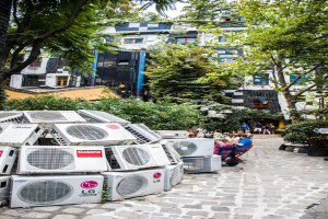 Luftkühler am Boden links unten, ein Weg nach rechts, Menschen auf Stühlen neben dem Weg sitzend, mit Gebäuden, Bäumen und Pflanzen oben.