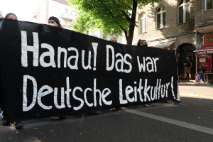 Eine Gruppe von Menschen geht eine Straße entlang und hält eine schwarze Fahne mit der Aufschrift "Hanau das war deutsche Leitkultur" hoch, mit Gebäuden, Bäumen und einem klaren blauen Himmel im Hintergrund.