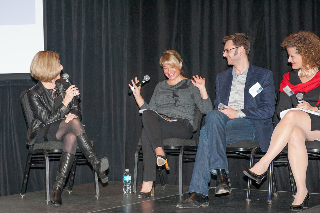 Ein Mann und drei Frauen sitzen auf Stühlen, halten jeweils ein Mikrofon und lächeln, mit einer Flasche, einem Bildschirm und Vorhängen im Hintergrund.