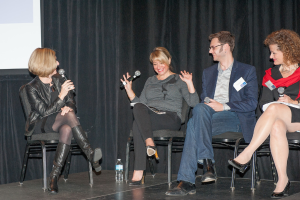 Ein Mann und drei Frauen sitzen auf Stühlen, halten jeweils ein Mikrofon und lächeln, mit einer Flasche, einem Bildschirm und Vorhängen im Hintergrund.