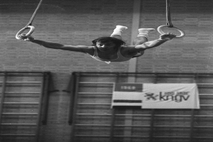 Ein Mann führt Turnübungen an den Ringen in einem Fitnessstudio durch, mit einer Wand und einem Banner mit Text im Hintergrund, dargestellt in Schwarz-Weiß.