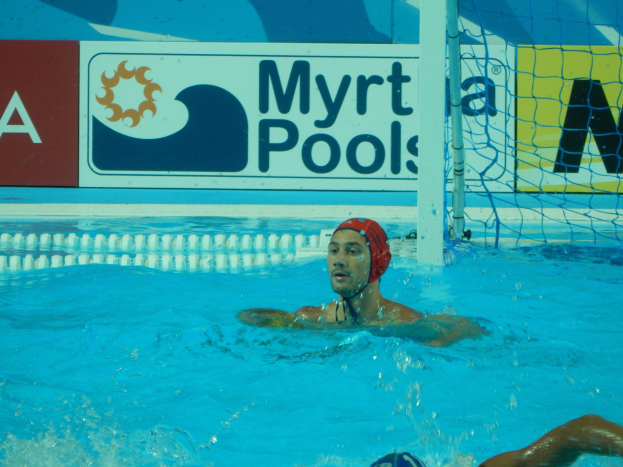 Wasserballspieler in Bewegung in einem Pool, tragend eine Kappe, mit einem Netz auf der rechten Seite und einer Werbetafel im Hintergrund.