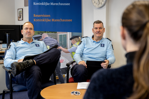 Zwei Polizeibeamte sitzen an einem Tisch und sprechen mit einer Frau; im Hintergrund sind eine Wandtafel, eine Uhr und Fotorahmen zu sehen, sowie ein weiterer Tisch mit Monitor, Tastatur und Maus links daneben.